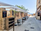 A charming outdoor cafe with a row of tables and chairs under large white umbrellas. The seating area is adjacent to a stone building with chalkboard menus displaying the day's specials, including dishes such as cannelloni. A small wooden shelf holds oranges and other items. The path leads to a scenic view of the sea in the background, with a tree providing some greenery. A few people are seen walking or standing nearby, enjoying the sunny day.