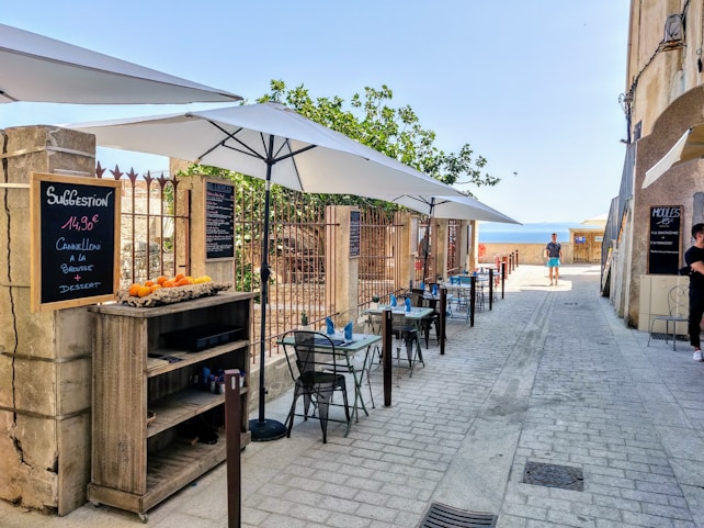 A charming outdoor cafe with a row of tables and chairs under large white umbrellas. The seating area is adjacent to a stone building with chalkboard menus displaying the day's specials, including dishes such as cannelloni. A small wooden shelf holds oranges and other items. The path leads to a scenic view of the sea in the background, with a tree providing some greenery. A few people are seen walking or standing nearby, enjoying the sunny day.