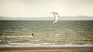 A person is kite surfing on a vast body of water with choppy waves. The sky is overcast, casting a subdued light on the scene. The kite is predominantly white with red accents, shaped like a parachute, and the surfer is wearing a wetsuit.