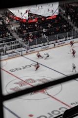 An indoor ice hockey game is taking place in an arena. The focus is on several players in action on the ice rink, with one player attempting to control the puck. The audience is visible in the stands, which are partially filled. A large overhead screen displays a live feed of the game, showing additional angles of the players. Advertising banners are positioned around the rink.