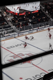 An indoor ice hockey game is taking place in an arena. The focus is on several players in action on the ice rink, with one player attempting to control the puck. The audience is visible in the stands, which are partially filled. A large overhead screen displays a live feed of the game, showing additional angles of the players. Advertising banners are positioned around the rink.