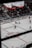 An indoor ice hockey game is taking place in an arena. The focus is on several players in action on the ice rink, with one player attempting to control the puck. The audience is visible in the stands, which are partially filled. A large overhead screen displays a live feed of the game, showing additional angles of the players. Advertising banners are positioned around the rink.