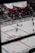 An indoor ice hockey game is taking place in an arena. The focus is on several players in action on the ice rink, with one player attempting to control the puck. The audience is visible in the stands, which are partially filled. A large overhead screen displays a live feed of the game, showing additional angles of the players. Advertising banners are positioned around the rink.