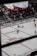 An indoor ice hockey game is taking place in an arena. The focus is on several players in action on the ice rink, with one player attempting to control the puck. The audience is visible in the stands, which are partially filled. A large overhead screen displays a live feed of the game, showing additional angles of the players. Advertising banners are positioned around the rink.