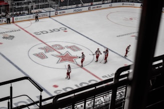 A group of ice hockey players in red uniforms are gathered on a rink, preparing for a game. The rink includes various advertisements along the boards and a large logo at center ice. The players are spread across the ice, each holding a hockey stick, and nearby officials in black and white stripes oversee the scene.