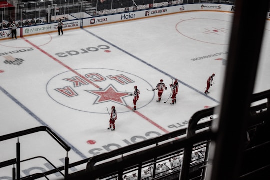 A group of ice hockey players in red uniforms are gathered on a rink, preparing for a game. The rink includes various advertisements along the boards and a large logo at center ice. The players are spread across the ice, each holding a hockey stick, and nearby officials in black and white stripes oversee the scene.