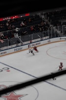 A hockey game in progress with players in action on the ice rink. The stadium seating shows a crowd watching the match, and advertisements line the rink's perimeter.