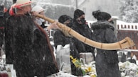 A person dressed in traditional clothing, wearing a hat and dark poncho, is playing a long wooden horn in a snowy outdoor setting. Several other people, also dressed warmly in coats and hats, stand nearby in the snow, suggesting they are part of an audience or event. Snow is falling heavily, covering the ground and surrounding objects.