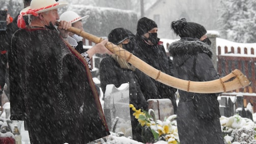 A person dressed in traditional clothing, wearing a hat and dark poncho, is playing a long wooden horn in a snowy outdoor setting. Several other people, also dressed warmly in coats and hats, stand nearby in the snow, suggesting they are part of an audience or event. Snow is falling heavily, covering the ground and surrounding objects.