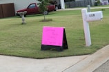 A residential neighborhood setting with a lawn and a pink sign indicating a garage sale. The sign is situated near a mailbox labeled '119' and a concrete pavement. A red pickup truck is parked in front of a house in the background.
