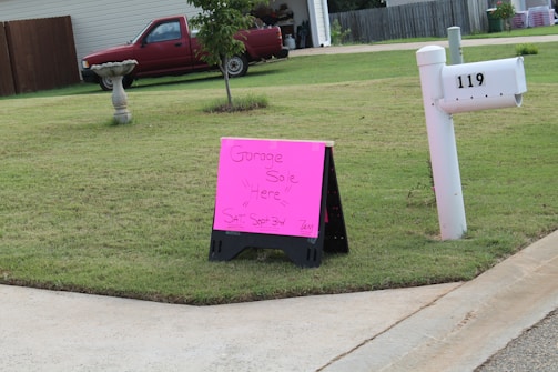 A residential neighborhood setting with a lawn and a pink sign indicating a garage sale. The sign is situated near a mailbox labeled '119' and a concrete pavement. A red pickup truck is parked in front of a house in the background.