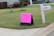 A residential neighborhood setting with a lawn and a pink sign indicating a garage sale. The sign is situated near a mailbox labeled '119' and a concrete pavement. A red pickup truck is parked in front of a house in the background.