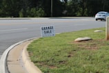 A roadside scene featuring a garage sale sign planted on a patch of green grass. The sign provides details about the sale, including the address and timings. In the background, a road with clear lane markings is visible, along with a white car parked or moving parallel to it. Trees line the other side of the road, and the environment appears to be a suburban or residential area.