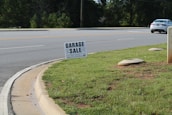 A roadside scene featuring a garage sale sign planted on a patch of green grass. The sign provides details about the sale, including the address and timings. In the background, a road with clear lane markings is visible, along with a white car parked or moving parallel to it. Trees line the other side of the road, and the environment appears to be a suburban or residential area.