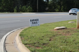 A roadside scene featuring a garage sale sign planted on a patch of green grass. The sign provides details about the sale, including the address and timings. In the background, a road with clear lane markings is visible, along with a white car parked or moving parallel to it. Trees line the other side of the road, and the environment appears to be a suburban or residential area.