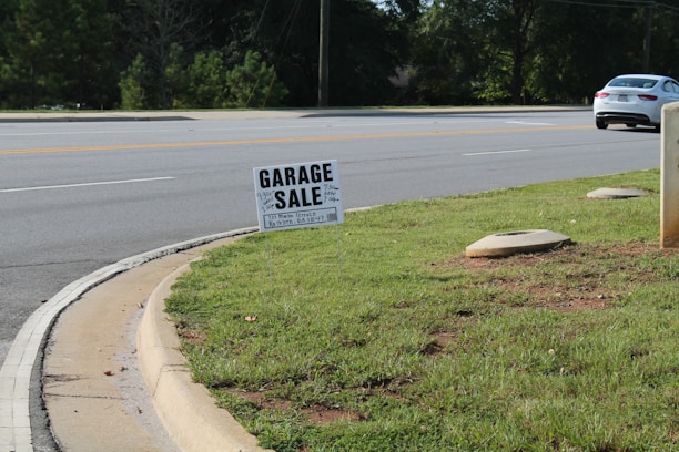 A roadside scene featuring a garage sale sign planted on a patch of green grass. The sign provides details about the sale, including the address and timings. In the background, a road with clear lane markings is visible, along with a white car parked or moving parallel to it. Trees line the other side of the road, and the environment appears to be a suburban or residential area.