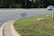 A roadside scene featuring a garage sale sign planted on a patch of green grass. The sign provides details about the sale, including the address and timings. In the background, a road with clear lane markings is visible, along with a white car parked or moving parallel to it. Trees line the other side of the road, and the environment appears to be a suburban or residential area.