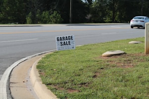 A roadside scene featuring a garage sale sign planted on a patch of green grass. The sign provides details about the sale, including the address and timings. In the background, a road with clear lane markings is visible, along with a white car parked or moving parallel to it. Trees line the other side of the road, and the environment appears to be a suburban or residential area.