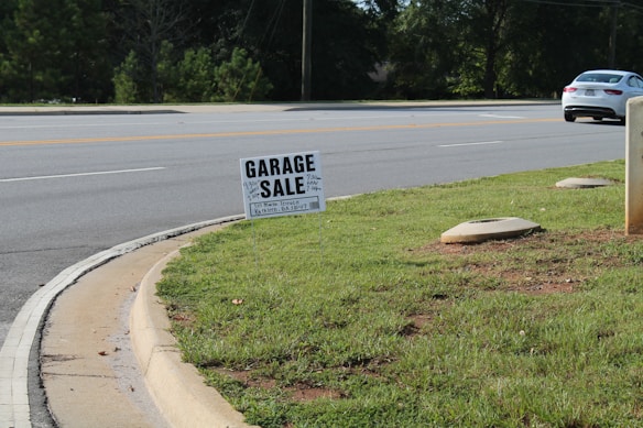 A roadside scene featuring a garage sale sign planted on a patch of green grass. The sign provides details about the sale, including the address and timings. In the background, a road with clear lane markings is visible, along with a white car parked or moving parallel to it. Trees line the other side of the road, and the environment appears to be a suburban or residential area.