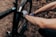 A person is wearing colorful gloves and repairing or adjusting a component on a black bicycle. The scene is set on a dirt path with scattered leaves, suggesting an outdoor environment.