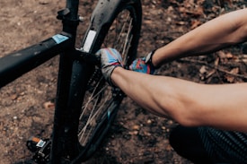 A person is wearing colorful gloves and repairing or adjusting a component on a black bicycle. The scene is set on a dirt path with scattered leaves, suggesting an outdoor environment.