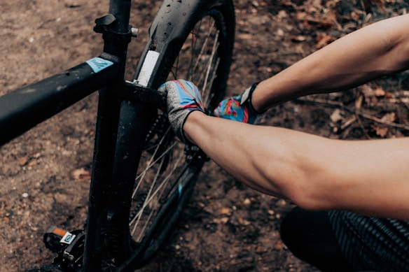 A person is wearing colorful gloves and repairing or adjusting a component on a black bicycle. The scene is set on a dirt path with scattered leaves, suggesting an outdoor environment.