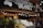 Fresh produce, including leafy greens and cauliflower, is displayed in wooden crates outside a market stall. The sign above reads 'South Melbourne Market Organics.' Ambient string lights hang, creating a warm and inviting atmosphere.