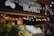 Fresh produce, including leafy greens and cauliflower, is displayed in wooden crates outside a market stall. The sign above reads 'South Melbourne Market Organics.' Ambient string lights hang, creating a warm and inviting atmosphere.