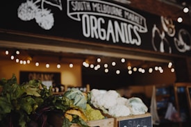 Fresh produce, including leafy greens and cauliflower, is displayed in wooden crates outside a market stall. The sign above reads 'South Melbourne Market Organics.' Ambient string lights hang, creating a warm and inviting atmosphere.