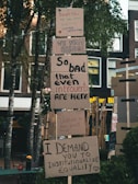 Protest signs are displayed on a stack of cardboard, with messages advocating for change and equality. The signs are placed outdoors in a setting with trees and buildings in the background. The messages convey a sense of urgency and unity.