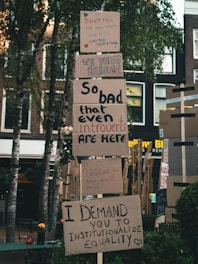 Protest signs are displayed on a stack of cardboard, with messages advocating for change and equality. The signs are placed outdoors in a setting with trees and buildings in the background. The messages convey a sense of urgency and unity.