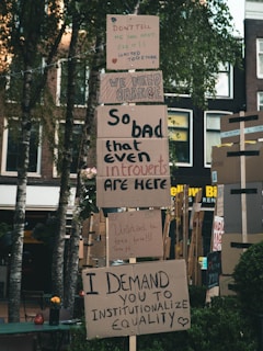 Protest signs are displayed on a stack of cardboard, with messages advocating for change and equality. The signs are placed outdoors in a setting with trees and buildings in the background. The messages convey a sense of urgency and unity.