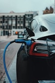 A sleek black electric car is parked on a city street, with a blue charging cable plugged into its port, indicating it is being charged. The background includes blurred buildings and street activity, suggesting an urban environment.