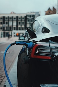A sleek black electric car is parked on a city street, with a blue charging cable plugged into its port, indicating it is being charged. The background includes blurred buildings and street activity, suggesting an urban environment.