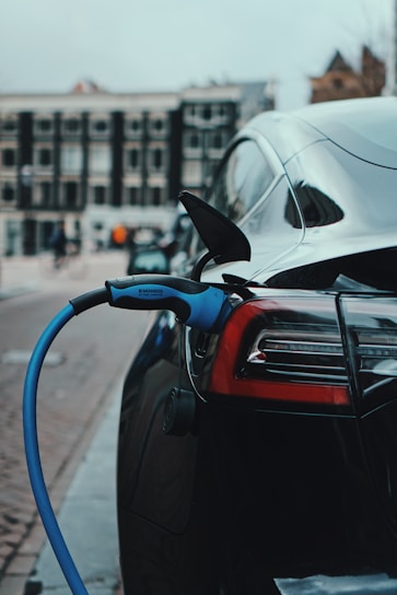 A sleek black electric car is parked on a city street, with a blue charging cable plugged into its port, indicating it is being charged. The background includes blurred buildings and street activity, suggesting an urban environment.