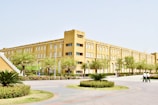 A large, multi-story academic building with a light brown facade is surrounded by landscaped greenery, including palm trees and neatly trimmed shrubs. A pathway leads to the entrance, and two people are walking nearby on a paved area. The architecture is simple and modern, featuring numerous windows and a staircase on one side.