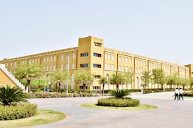 A large, multi-story academic building with a light brown facade is surrounded by landscaped greenery, including palm trees and neatly trimmed shrubs. A pathway leads to the entrance, and two people are walking nearby on a paved area. The architecture is simple and modern, featuring numerous windows and a staircase on one side.