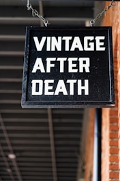 A black rectangular sign with bold white letters hangs from a chain, displaying the words 'VINTAGE AFTER DEATH'. The background features a brick wall on the right side and a ceiling with exposed beams.