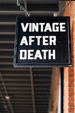 A black rectangular sign with bold white letters hangs from a chain, displaying the words 'VINTAGE AFTER DEATH'. The background features a brick wall on the right side and a ceiling with exposed beams.