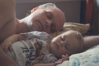 A man and a child are peacefully sleeping together, resting on a bed with soft light illuminating their faces. The child is wearing a white shirt with colorful designs while the man's arm is gently wrapped around the child, creating a sense of comfort and safety.