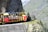 A colorful open-air train travels along a narrow mountain track with lush greenery and rocky cliffs in the background. Several passengers are seated in the compartment, enjoying the scenic view.