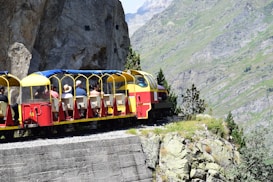 A colorful open-air train travels along a narrow mountain track with lush greenery and rocky cliffs in the background. Several passengers are seated in the compartment, enjoying the scenic view.