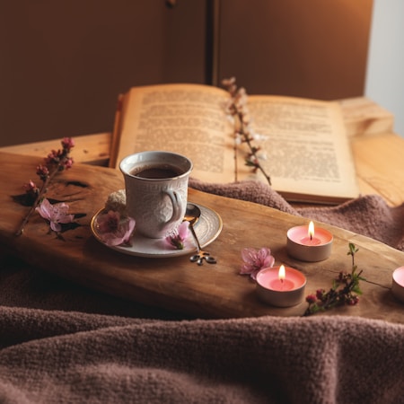A serene scene features an open book resting on a wooden surface surrounded by decorative elements. A white porcelain cup filled with coffee sits on a saucer adorned with pink blossoms. Several lit tea candles add a soft glow, and the setup is complemented by a plush brown blanket, creating a cozy atmosphere.