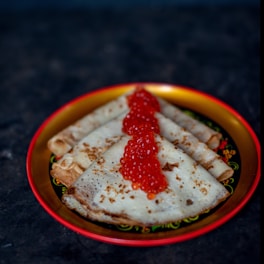 A decorative plate holds several folded crepes with golden brown spots. A generous amount of vibrant red caviar is placed on top of the crepes, adding a touch of elegance to the dish. The plate has a colorful border, enhancing the visual appeal against the dark background.