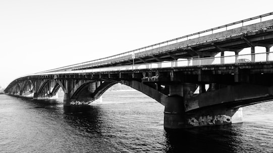A black and white image of a large concrete bridge spanning across a body of water. The bridge features multiple arches and railings, with some graffiti visible on the lower sections of the structure. The water below is calm and reflects the bridge.