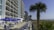 A tall, multi-story beachfront hotel is visible with several balconies facing the ocean. In front of the building, there are numerous patio tables and chairs with blue umbrellas, next to a pool. A palm tree is prominently displayed on the right side, with more palm trees in the background along the shoreline. The sky is clear and blue.