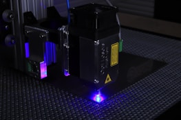 A close-up of a laser engraving machine working on a textured surface. The machine's laser emits a bright blue light, illuminating the surrounding area. The metal parts of the machine have labels and warning signs, and there are several metallic and plastic components visible.