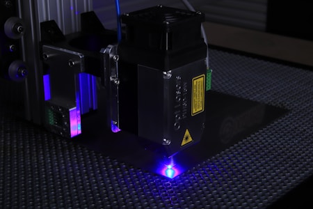 A close-up of a laser engraving machine working on a textured surface. The machine's laser emits a bright blue light, illuminating the surrounding area. The metal parts of the machine have labels and warning signs, and there are several metallic and plastic components visible.