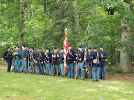 A group of people dressed in historical military uniforms are standing in a forested area. They are wearing blue jackets and pants, holding rifles, and carrying flags, which suggests a Civil War reenactment. The setting is lush with green trees and grass.