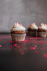 A close-up of a chocolate cupcake topped with a swirl of white frosting and sprinkled with red crumbs. In the background, there are more cupcakes, slightly out of focus. The surface is dusted with scattered red crumbs, creating a visually appealing texture against the dark backdrop.